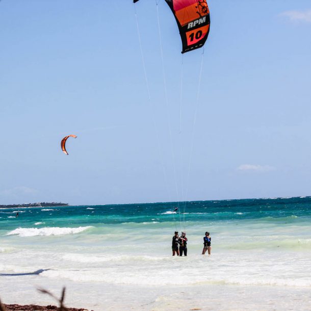 2 women and their instructor learning to fly a kite for the first time in Tulum Mexico Camp. White sand and clear waters as their landscape and another kitesurfers riding further upwind.