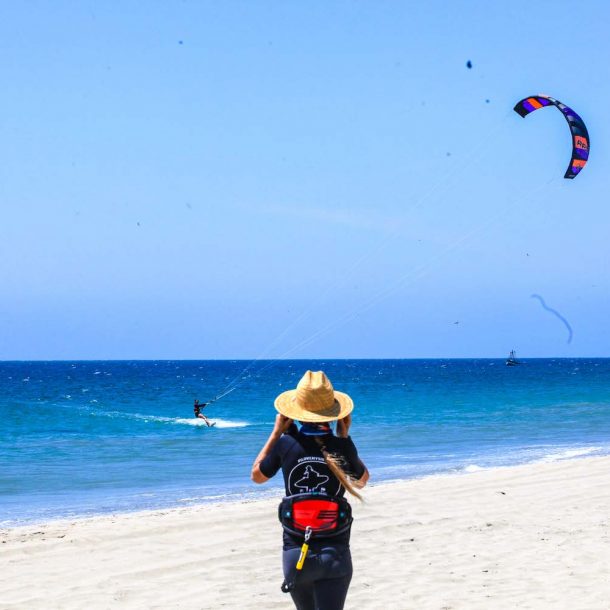 Woman taking a photo of a female kitesurfer that is in the water riding, standing on a beautiful sandy beach and turquoise blue sea during Women Kiteboarding Camp